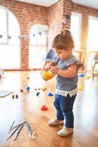 toddler girl holding a play cup in a room with various toys on the floor
